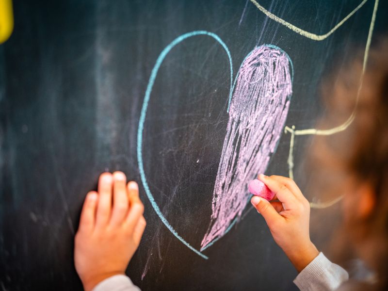 Kind malt mit rosa Kreide ein Herz an einer Tafel aus