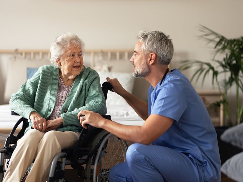 Jüngerer Pfleger kniet und spricht mit älteren Dame im Rollstuhl